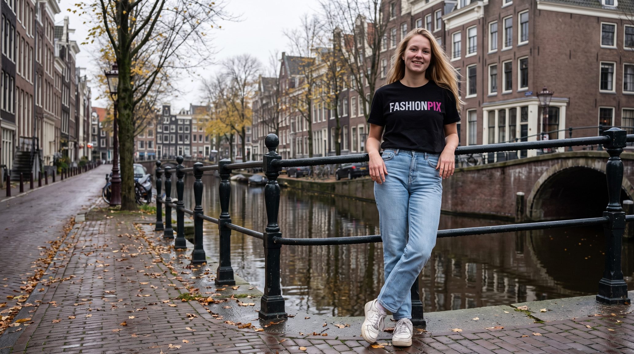 Jong blond model in het zwarte FASHIONPIX-T-shirt en een lichte jeans leunt glimlachend tegen de reling aan een Amsterdamse gracht met een bruggetje